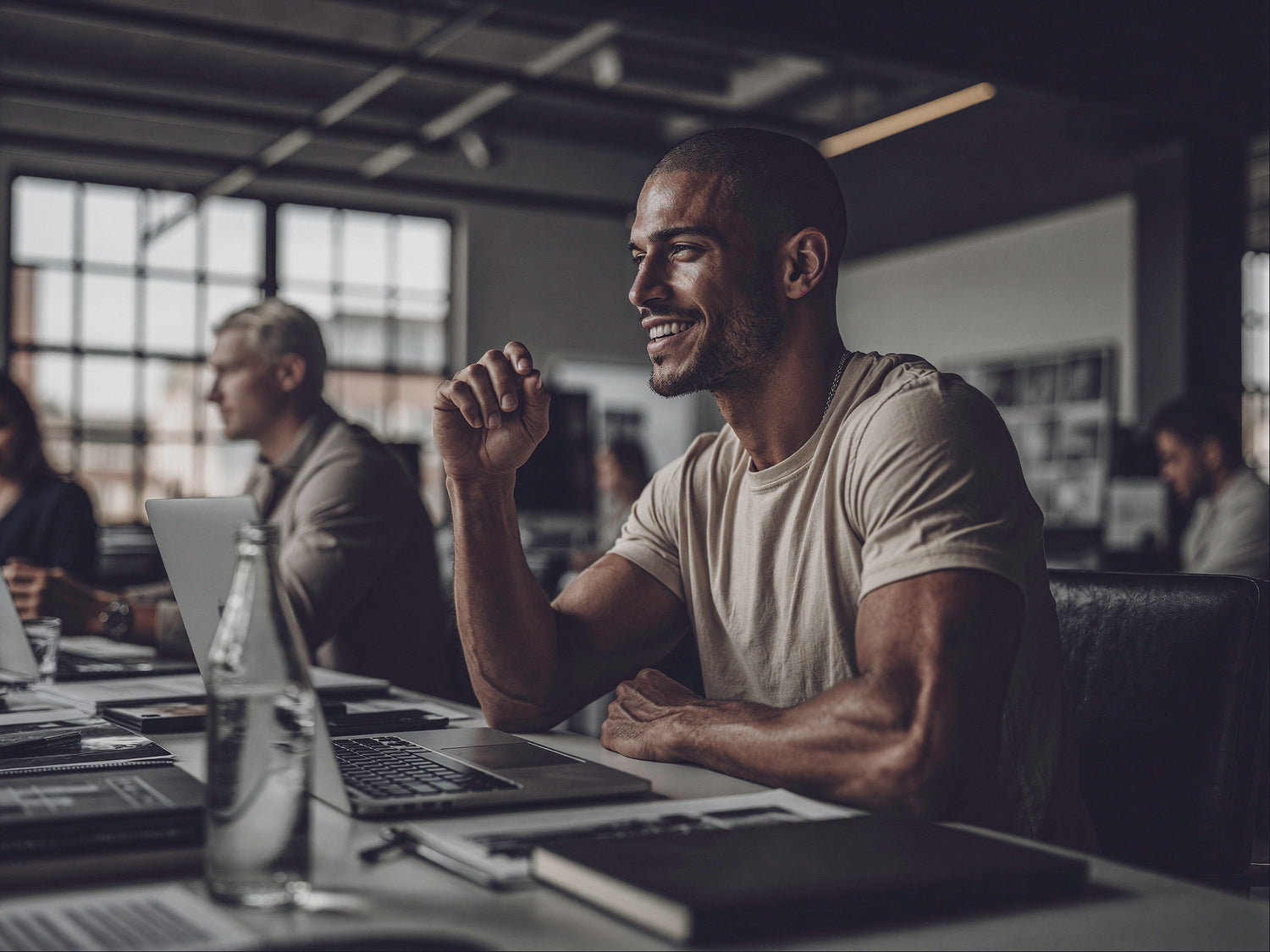 Muscular man with a buzz cut smiling during a modern office meeting, seated at a conference table with laptops and notebooks in a warm industrial workspace, representing energy, focus, and productivity after recovery support strips.