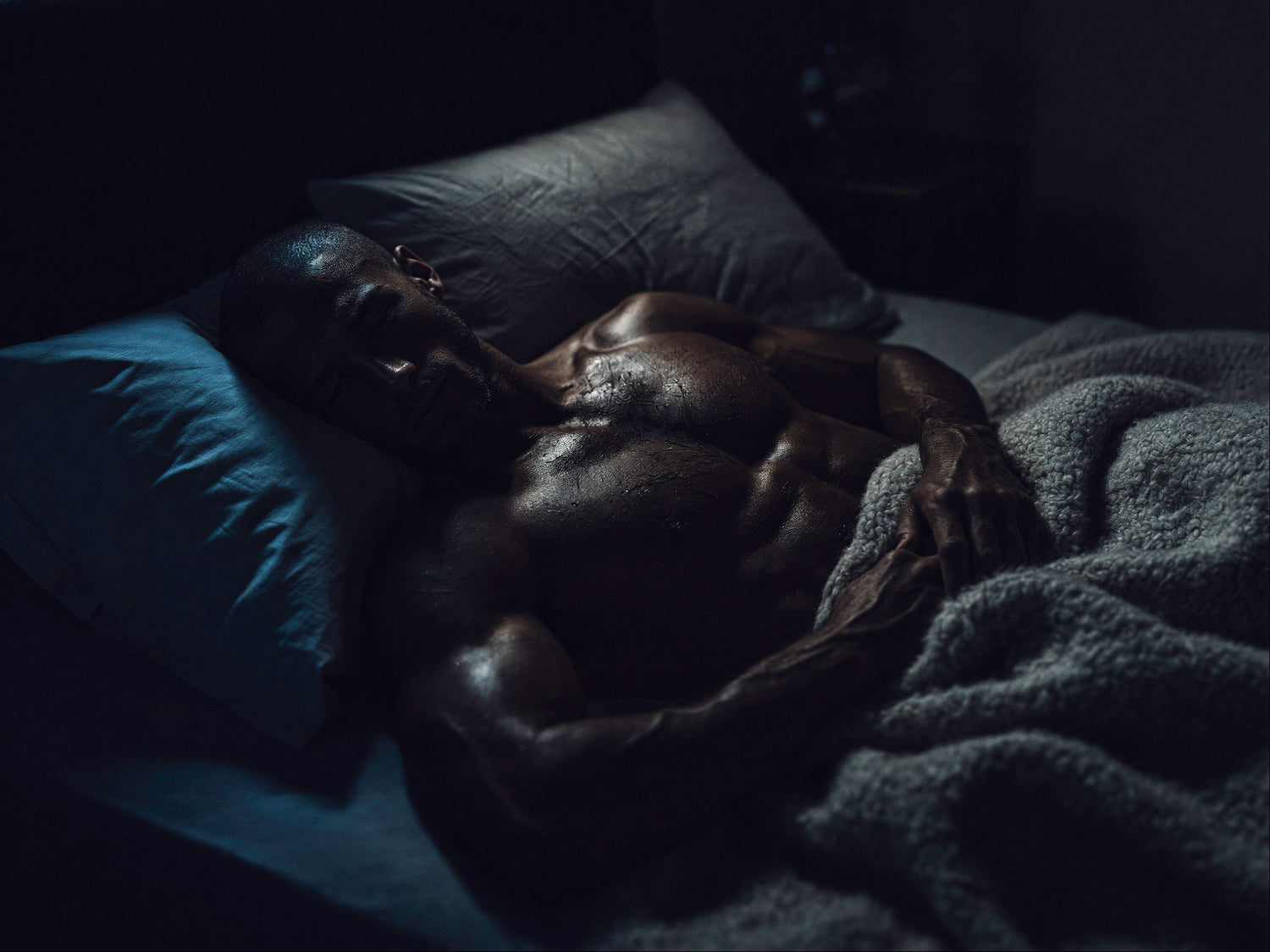 Shirtless muscular man peacefully sleeping in a dark bedroom under a textured blanket with cool blue nighttime lighting, representing deep sleep, nighttime recovery, and calming wellness support.
