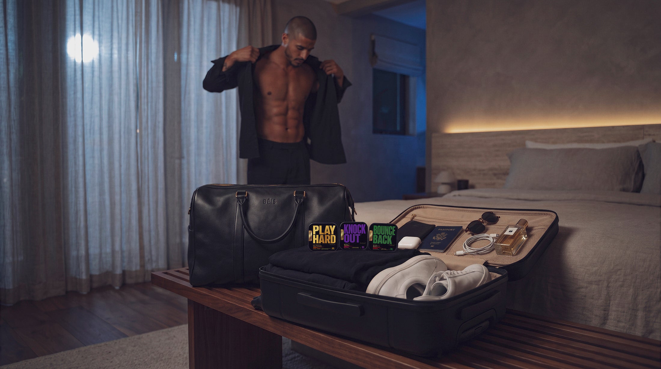 Three ['WELL]™ oral strip tins—PLAY HARD™, KNOCK OUT™, and BOUNCE BACK™—displayed in an open suitcase with weekend essentials while a man gets dressed in the background of a modern bedroom.